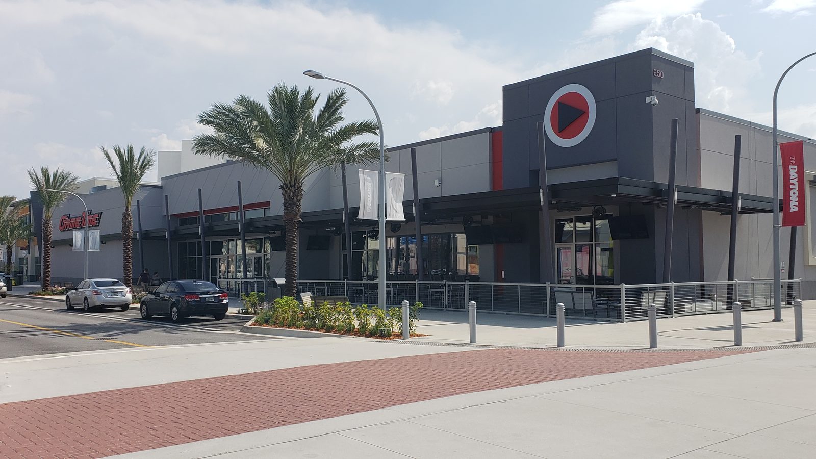 The outside of GameTime Daytona: a dark gray storefront with the play-button logo above the entrance, palm trees and a fenced patio along the sidewalk