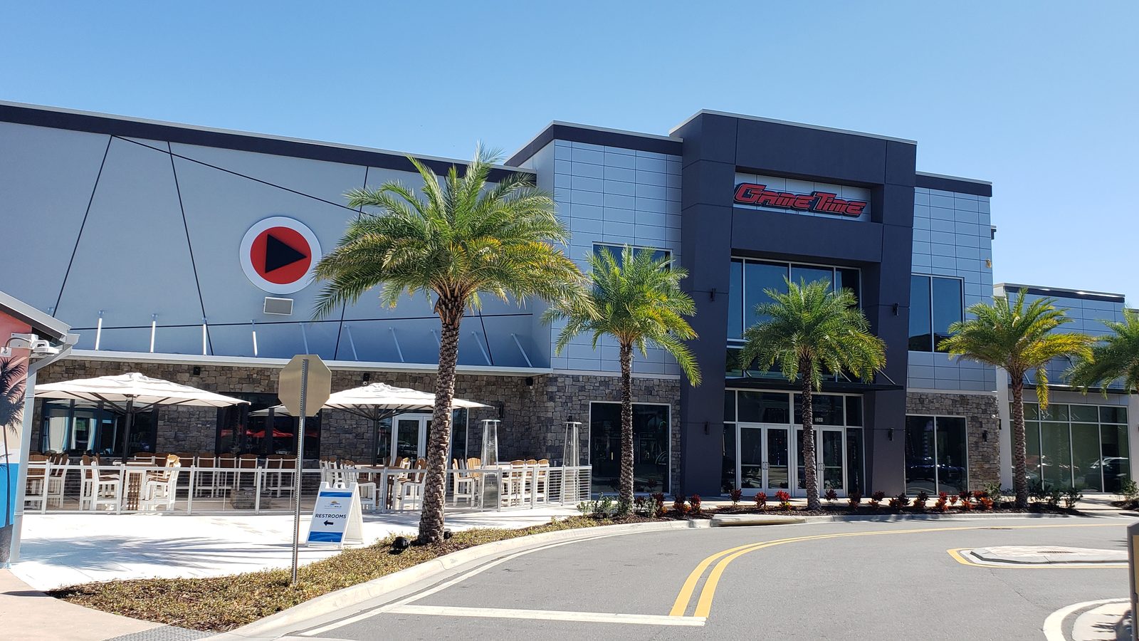 The outside of GameTime Kissimmee: a gray two-story front with the red GameTime sign and play-button logo, palm trees along the walk and a covered patio with umbrellas at the left