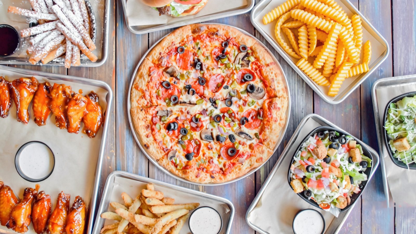 An overhead spread of GameTime food: a supreme pizza, jumbo wings, crinkle fries, a salad, fried pickle sticks and powdered churros
