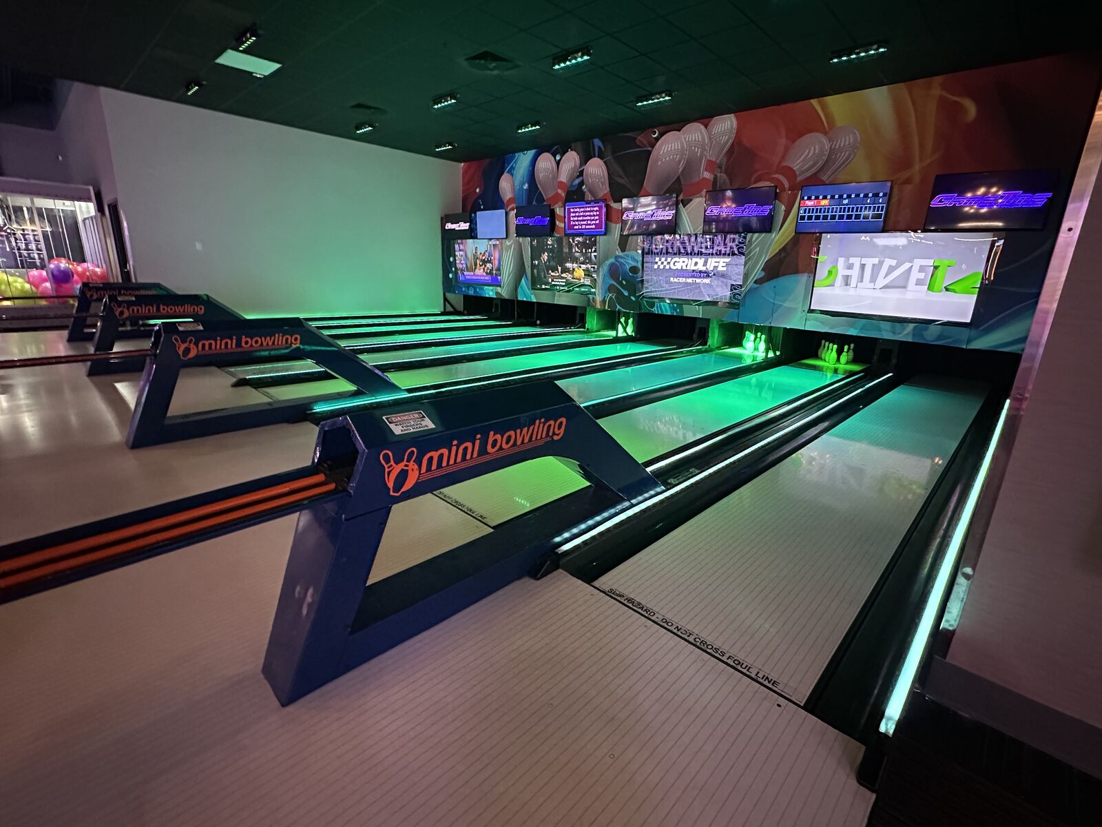 The mini bowling lanes at GameTime Kissimmee from the ball return, the lanes lit green beneath a wall of screens