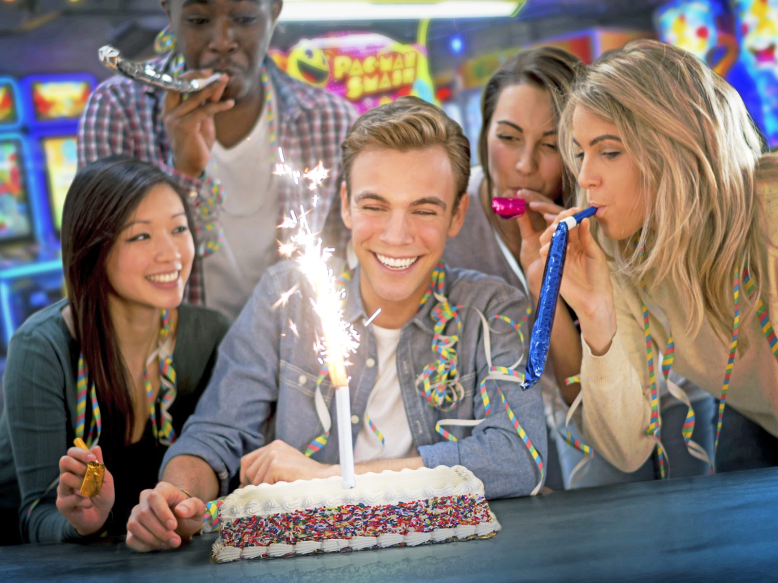Friends at a GameTime birthday party leaning in over a sprinkle-covered cake with a lit sparkler, party horns and streamers in hand, arcade games behind them