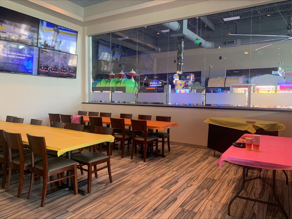 The event space at GameTime Kissimmee: tables set with yellow and pink cloths beside a glass wall over the game floor, with screens above