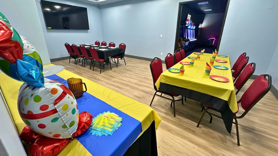 A private party room at GameTime Ocoee set for a birthday, with yellow-covered tables, rainbow plates and cups, red chairs, balloons, and a doorway through to the game floor