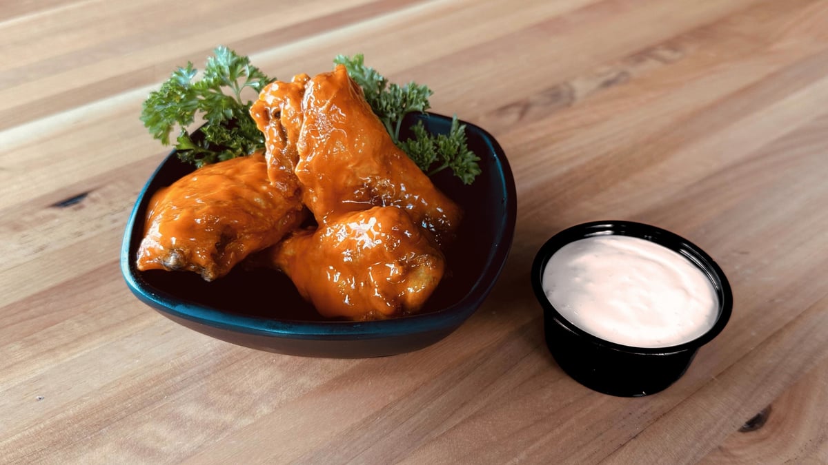 Three jumbo wings in an orange wing sauce, in a dark bowl with parsley, beside a cup of white dipping sauce on a wooden table