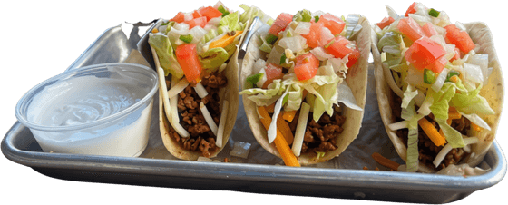 Three beef tacos with lettuce, tomato and cheese on a metal tray, with a cup of white sauce